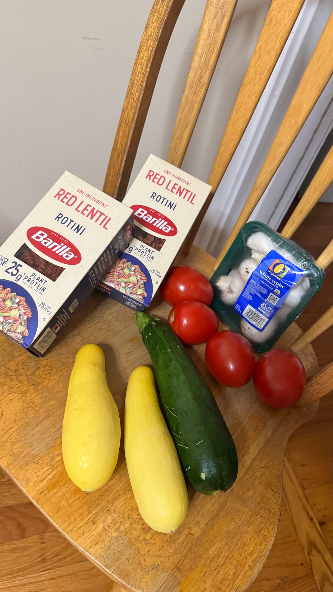 Red lentil rotini boxes, zucchini, yellow squash, Roma tomatoes, and white mushrooms on a wooden chair
